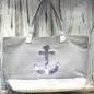 Preview: Graue Shoppertasche mit silbernem Pailletten-Anker, weißem Boden mit Silberstreifen und grauen Kunstleder-Griffen vor rustikalem Holzhintergrund.